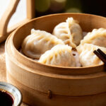 Raviolis chinois vapeur frais disposés dans un panier en bambou, légèrement brillants avec des plis délicats, accompagnés de baguettes et d’un petit bol de sauce soja avec graines de sésame et piment.