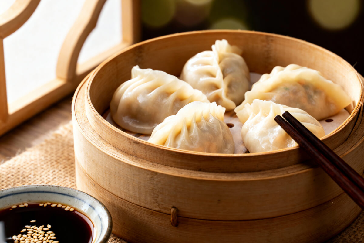 Raviolis chinois vapeur frais disposés dans un panier en bambou, légèrement brillants avec des plis délicats, accompagnés de baguettes et d’un petit bol de sauce soja avec graines de sésame et piment.